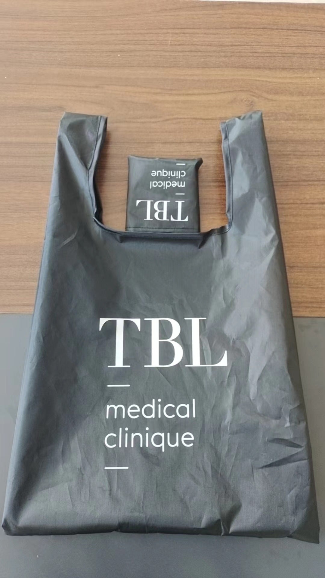 Black tank-top foldable shopping bag printed with TBL medical clinique logo, shown folded and ready for custom branding