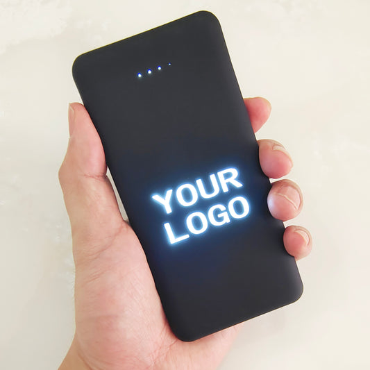 Black rectangular power bank with glowing LED logo panel and multiple USB ports on a dark surface